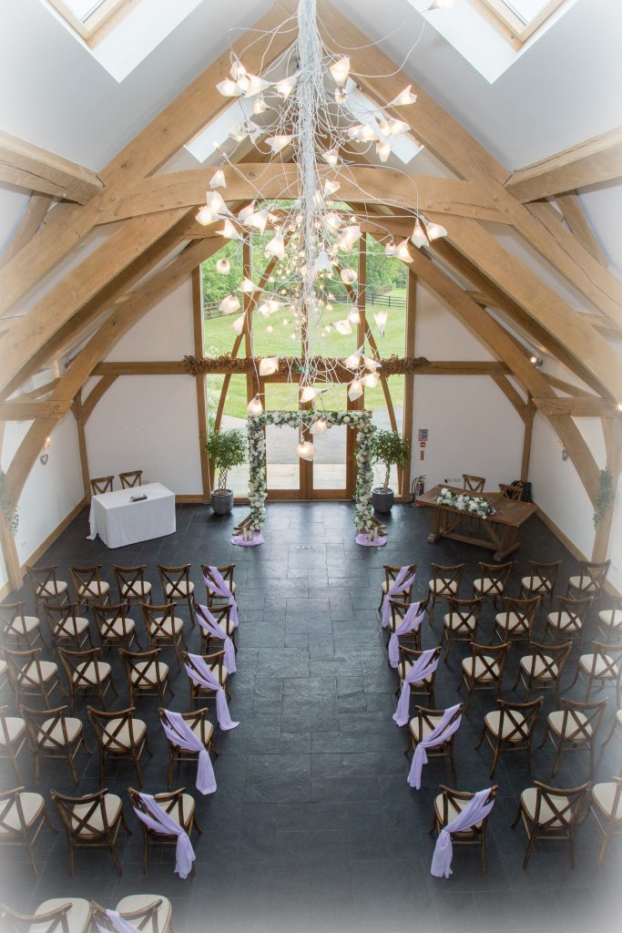 Mythe barn ceremony space with lilac chiffon chair drapes and rustic arch, styled with full ivory and lilac flowers. Match Florals on the register table