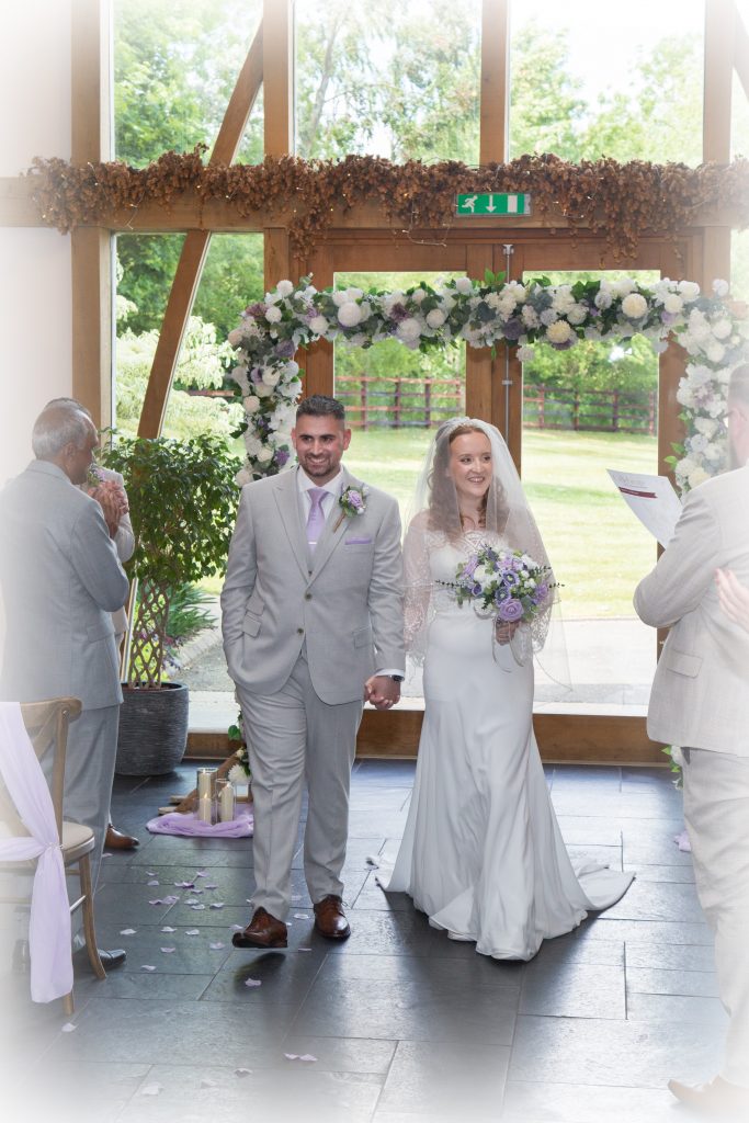 Bride and groom walking up the aisle after getting married with an arch covered in ivory and lilac flowers in the background